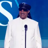Niewidomy piosenkarz Stevie Wonder w białym garniturze i czarnej czapce, przemawia do mikrofonu na tle niebieskiej sceny z logo CBS.