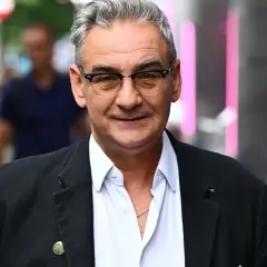 Robert Gonera w okularach, w czarnej marynarce i białej koszuli, uśmiecha się. W tle widać ludzi i różowe światła.