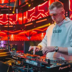 DJ w białej bluzie z kapturem gra na konsoli DJ-skiej na tle czerwonych świateł scenicznych.