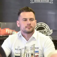 Grzegorz Collins, młody mężczyzna w białej koszuli, z tatuażem na szyi, siedzi przy stole. W tle widoczny pomarańczowy but sportowy.