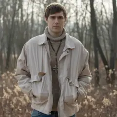 Sebastian Keller w beżowej kurtce i z krzyżem na szyi, w tle las.