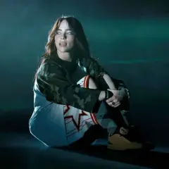 Billie Eilish siedzi na scenie, wpatrzona w dal. Jej stylizacja i mroczny klimat sugerują, że zaraz usłyszymy fajne piosenki.