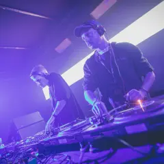Dwóch DJ-ów w słuchawkach tworzy muzykę techno. Jeden z nich ma na szyi identyfikator "ARTIST".