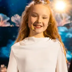 Uśmiechnięta dziewczynka z błyszczącym makijażem na oczach, w białej sukience, gotowa do występu na Eurowizji Junior Polska.