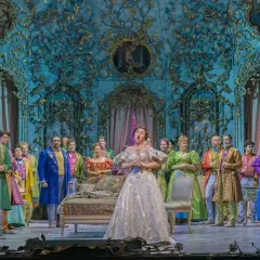 Primadonna w lśniącej sukni śpiewa na scenie operowej, otoczona przez barwnie ubranych artystów.
