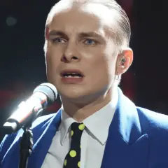 Ralph Kaminski, w niebieskiej marynarce i krawacie w grochy, śpiewa do mikrofonu. Jego partner, wokalista, jest w centrum uwagi.