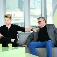 Kris Cugowski i jego syn rozmawiają na kanapie. Młodszy z nich, z blond włosami, gestykuluje, starszy, w okularach, z uśmiechem słucha.