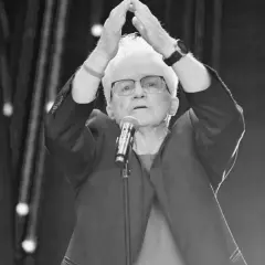 Starsza pani z białymi włosami, w okularach, z rękami uniesionymi nad głową, przed mikrofonem. To może być Jolka podczas występu.