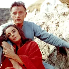 Elizabeth Taylor, żona Richarda Burtona, opiera głowę na jego ramieniu, otulona czerwoną narzutą, siedząc na kamieniach.