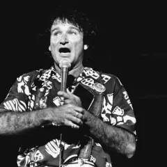 Robin Williams, w hawajskiej koszuli, śpiewa do mikrofonu, trzymając w dłoni notes.
