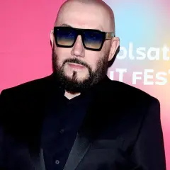 Skawiński w czarnym garniturze i okularach przeciwsłonecznych pozuje na tle różowego banera z logo Polsatu.