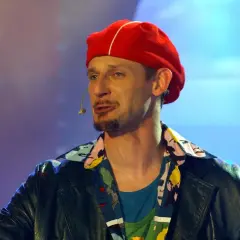 Michał Wójcik w czerwonej beretce i kolorowej koszuli, z mikrofonem przy ustach, na tle niebiesko-fioletowego światła.