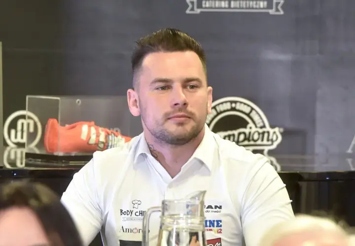 Grzegorz Collins, młody mężczyzna w białej koszuli, z tatuażem na szyi, siedzi przy stole. W tle widoczny pomarańczowy but sportowy.
