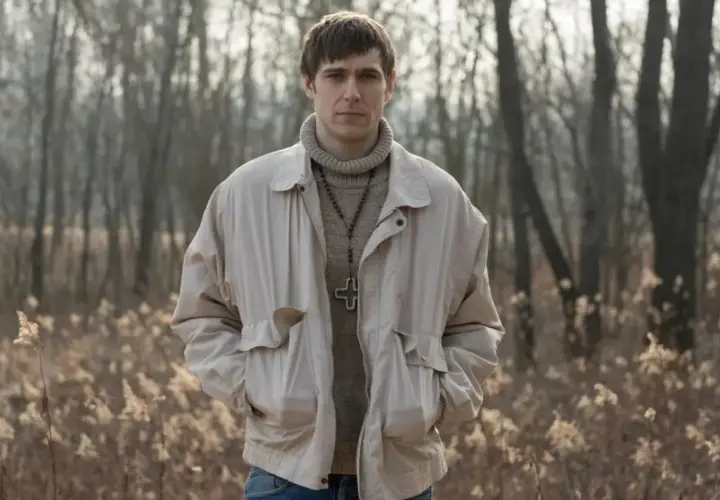 Sebastian Keller w beżowej kurtce i z krzyżem na szyi, w tle las.