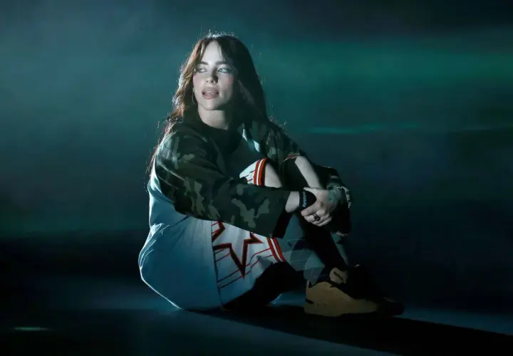Billie Eilish siedzi na scenie, wpatrzona w dal. Jej stylizacja i mroczny klimat sugerują, że zaraz usłyszymy fajne piosenki.
