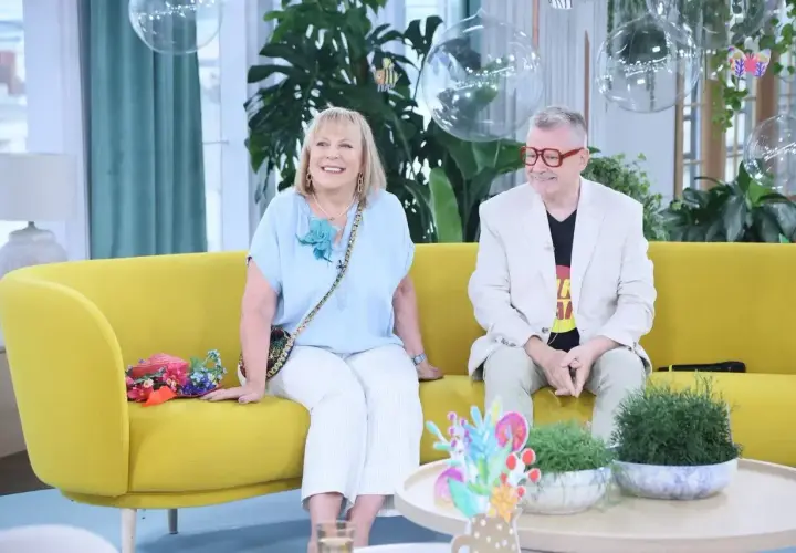 Uśmiechnięta para siedzi na żółtej kanapie. Pan Tik Tak w jasnej marynarce i kolorowym T-shircie.