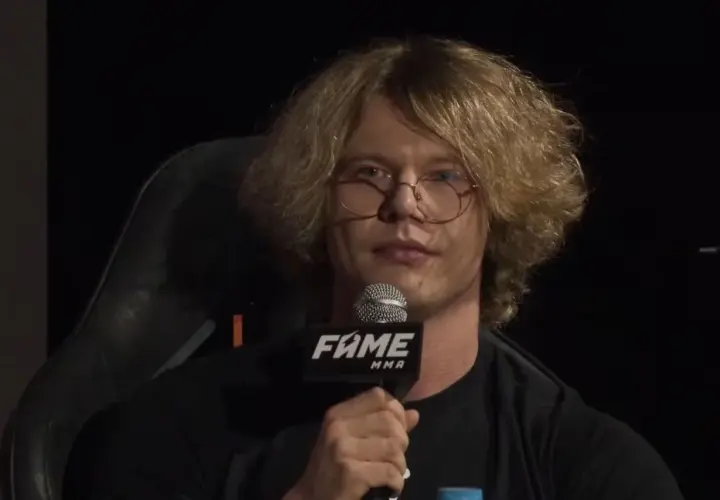 Natan Marcon z burzą blond włosów i okularami, trzymający mikrofon FAME MMA. Jego wiek jest trudny do określenia, ale wygląda na młodego mężczyznę.