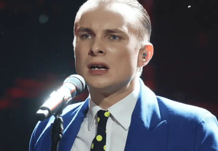 Ralph Kaminski, w niebieskiej marynarce i krawacie w grochy, śpiewa do mikrofonu. Jego partner, wokalista, jest w centrum uwagi.