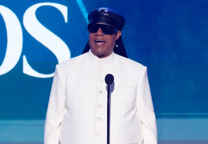 Niewidomy piosenkarz Stevie Wonder w białym garniturze i czarnej czapce, przemawia do mikrofonu na tle niebieskiej sceny z logo CBS.