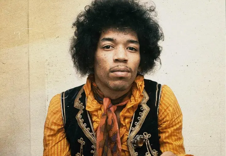 Jimi Hendrix w pomarańczowej koszuli i kamizelce, z charakterystyczną fryzurą. W tle subtelna, lekko zatarte tło, jakby w **purple haze**.