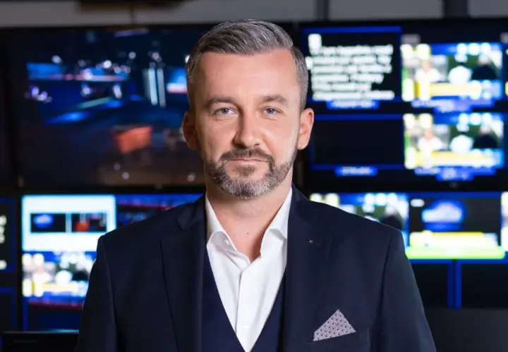 Krzysztof Skórzyński, elegancki mężczyzna w garniturze, stoi na tle ekranów. Ile ma lat? Tego nie wiemy, ale wygląda na doświadczonego dziennikarza.