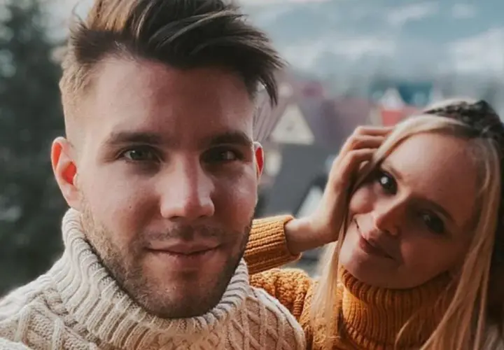 Olga Kalicka i jej partner w ciepłych swetrach, uśmiechnięci na tle górskiego krajobrazu.