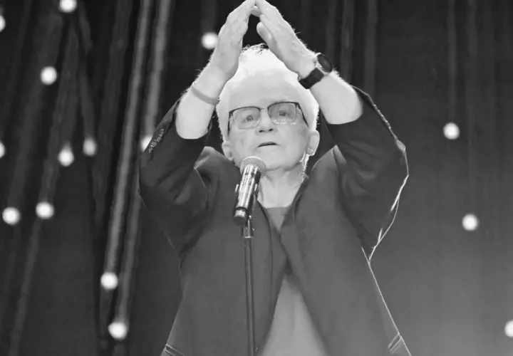 Starsza pani z białymi włosami, w okularach, z rękami uniesionymi nad głową, przed mikrofonem. To może być Jolka podczas występu.
