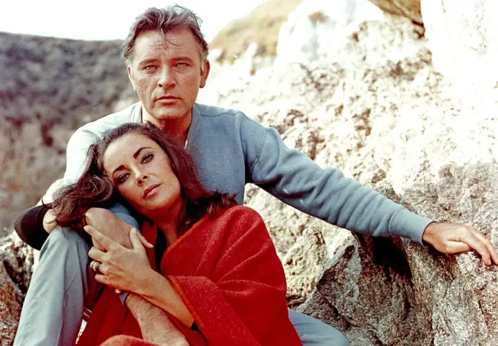 Elizabeth Taylor, żona Richarda Burtona, opiera głowę na jego ramieniu, otulona czerwoną narzutą, siedząc na kamieniach.