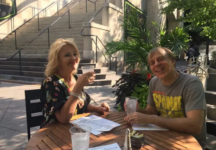 Tom Logan, mąż Majki Jeżowskiej, uśmiecha się do zdjęcia, siedząc przy stoliku na zewnątrz z drinkiem.