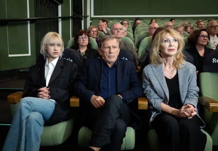 Małgorzata Englert, w wieku, siedzi w pierwszym rzędzie obok innych osób, wpatrując się w dal.