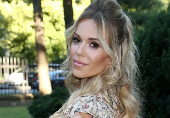 Doda IQ, blondynka w kwiecistej sukience, z burzą loków, uśmiecha się do obiektywu na tle zieleni.