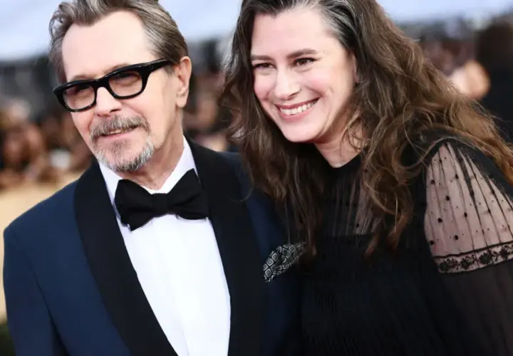 Gary Oldman z żoną, oboje uśmiechnięci, on w garniturze i okularach, ona w czarnej sukience.