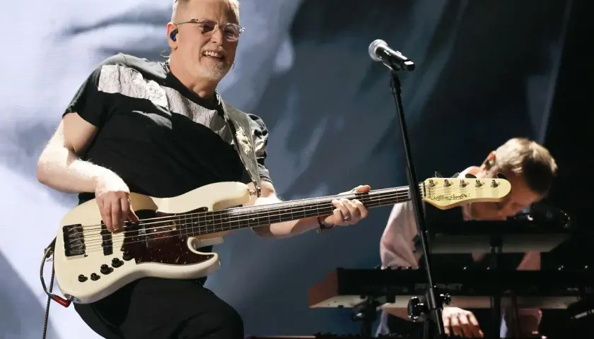Muzyk gra na białej gitarze basowej, tworząc swoje opus magnum. W tle klawiszowiec pochyla się nad instrumentem.