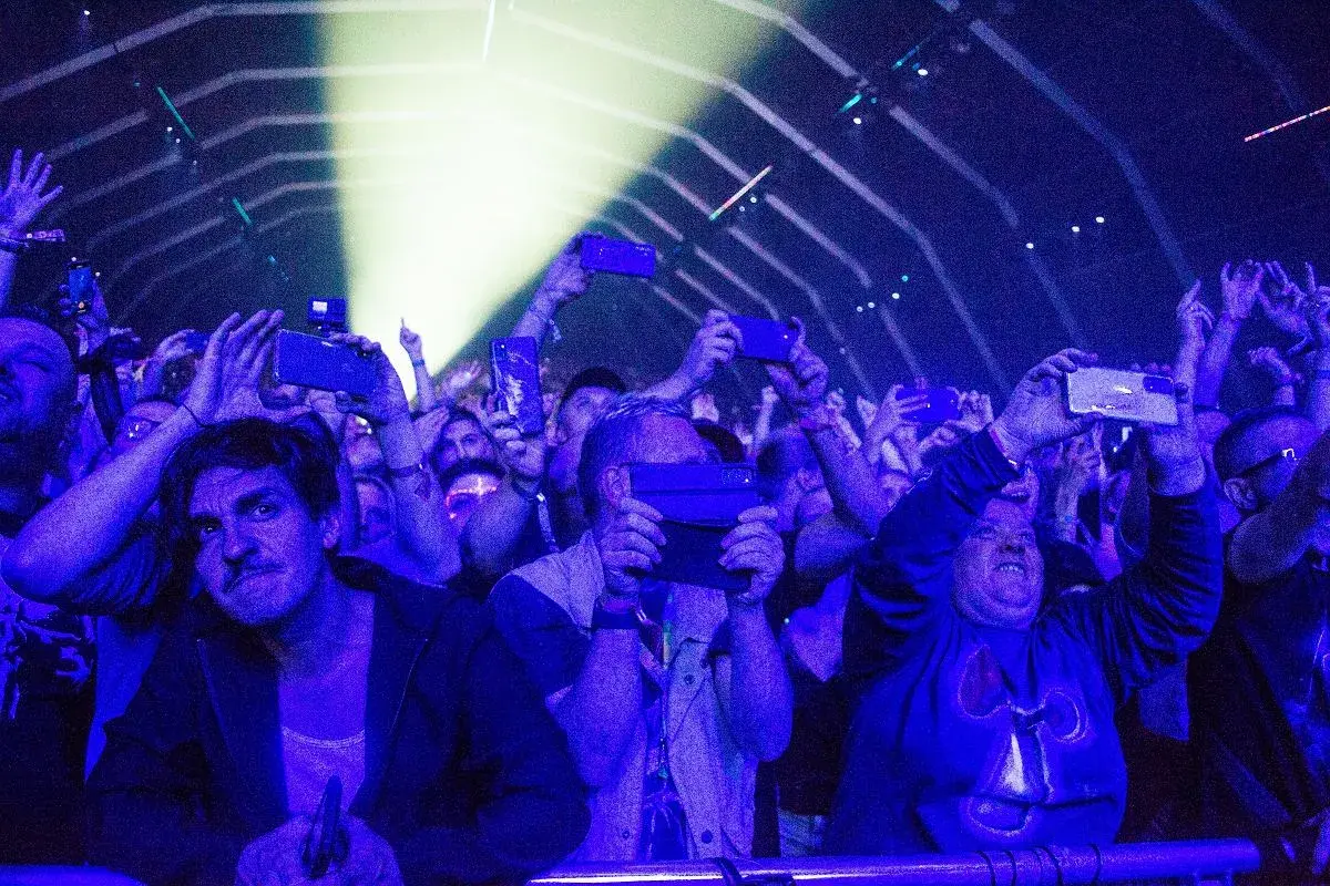 Tłum na fest festiwalu nagrywa koncert telefonami. Światła sceny oświetlają twarze ludzi.