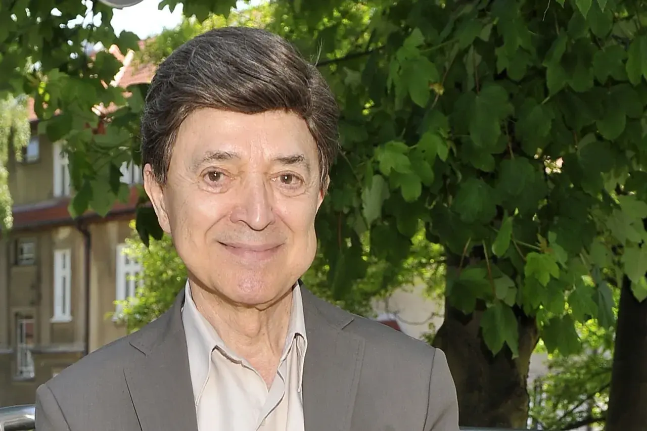 Uśmiechnięty Jerzy Połomski, w szarym garniturze i jasnej koszuli, na tle zieleni. Czyżby szukał partnera do wspólnego śpiewania?