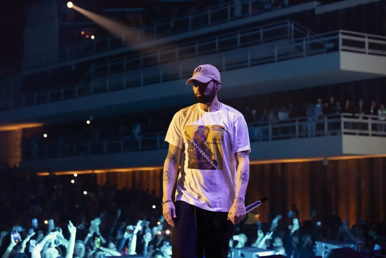 Eminem w czapce i białej koszulce z grafiką, na koncercie przed publicznością.