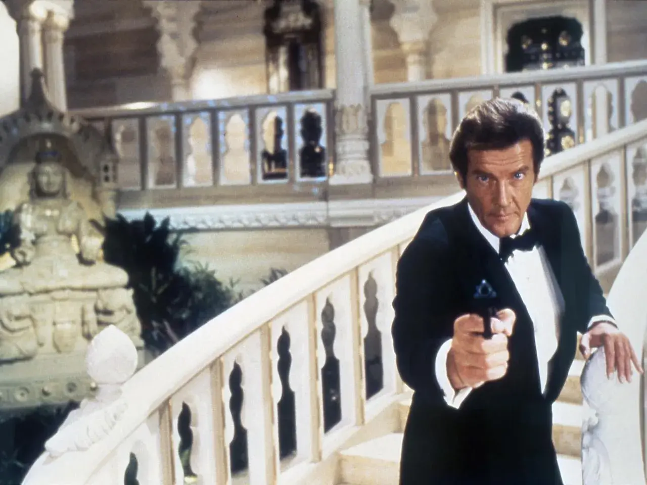 Roger Moore w smokingu z pistoletem, jakby szukał swojej żony w egzotycznym pałacu.