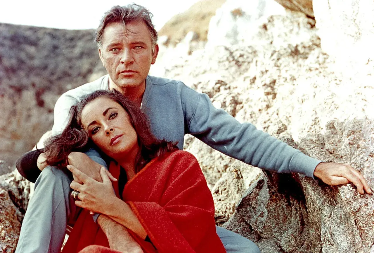 Elizabeth Taylor, żona Richarda Burtona, opiera głowę na jego ramieniu, otulona czerwoną narzutą, siedząc na kamieniach.