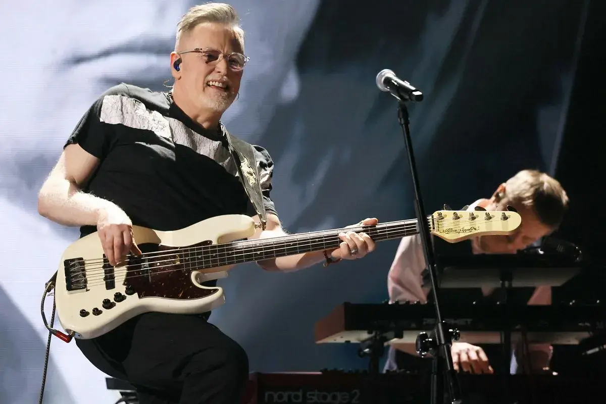 Muzyk gra na białej gitarze basowej, tworząc swoje opus magnum. W tle klawiszowiec pochyla się nad instrumentem.