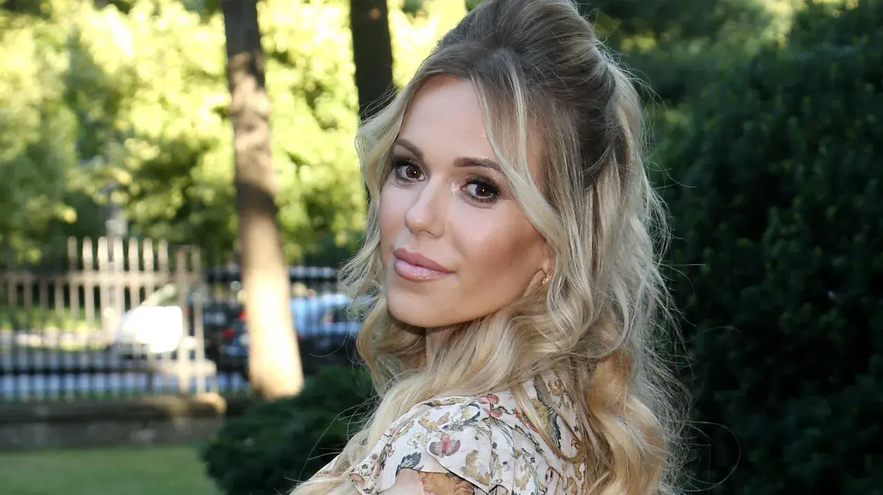 Doda IQ, blondynka w kwiecistej sukience, z burzą loków, uśmiecha się do obiektywu na tle zieleni.