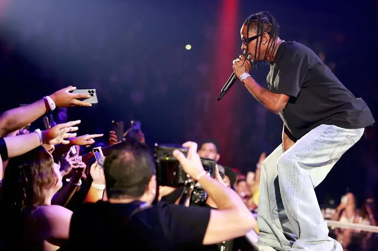 Travis Scott na koncercie, śpiewa do mikrofonu, a fani z telefonami próbują go nagrać.