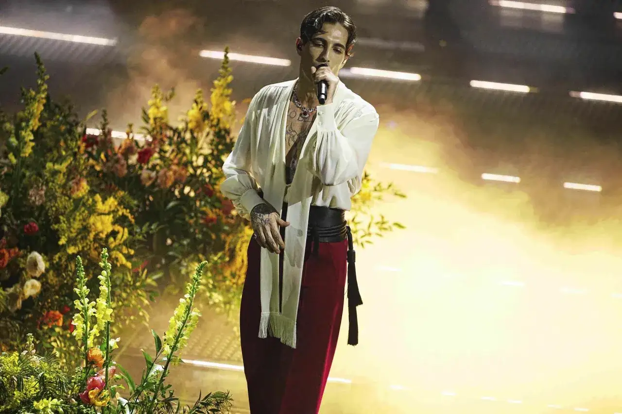 Damiano David na koncercie, w białej koszuli i czerwonych spodniach, śpiewa na tle bujnych kwiatów i dymu.