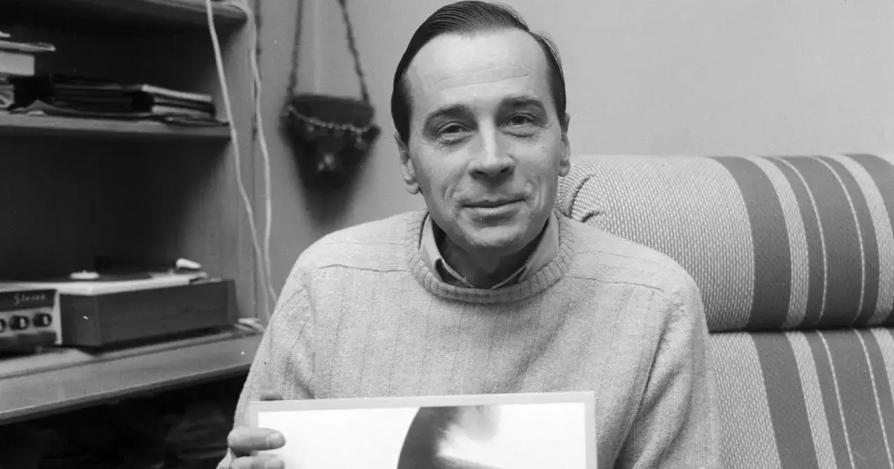 Edmund Fetting z partnerem, siedzący na kanapie, trzymający zdjęcie. W tle półka z książkami i gramofon.