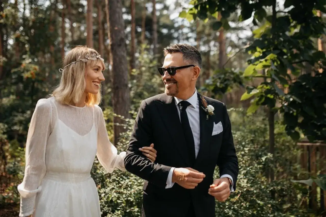 Krzysztof Ibisz z żoną, szczęśliwi idą przez las. Ona w białej sukni i wianku, on w eleganckim garniturze.