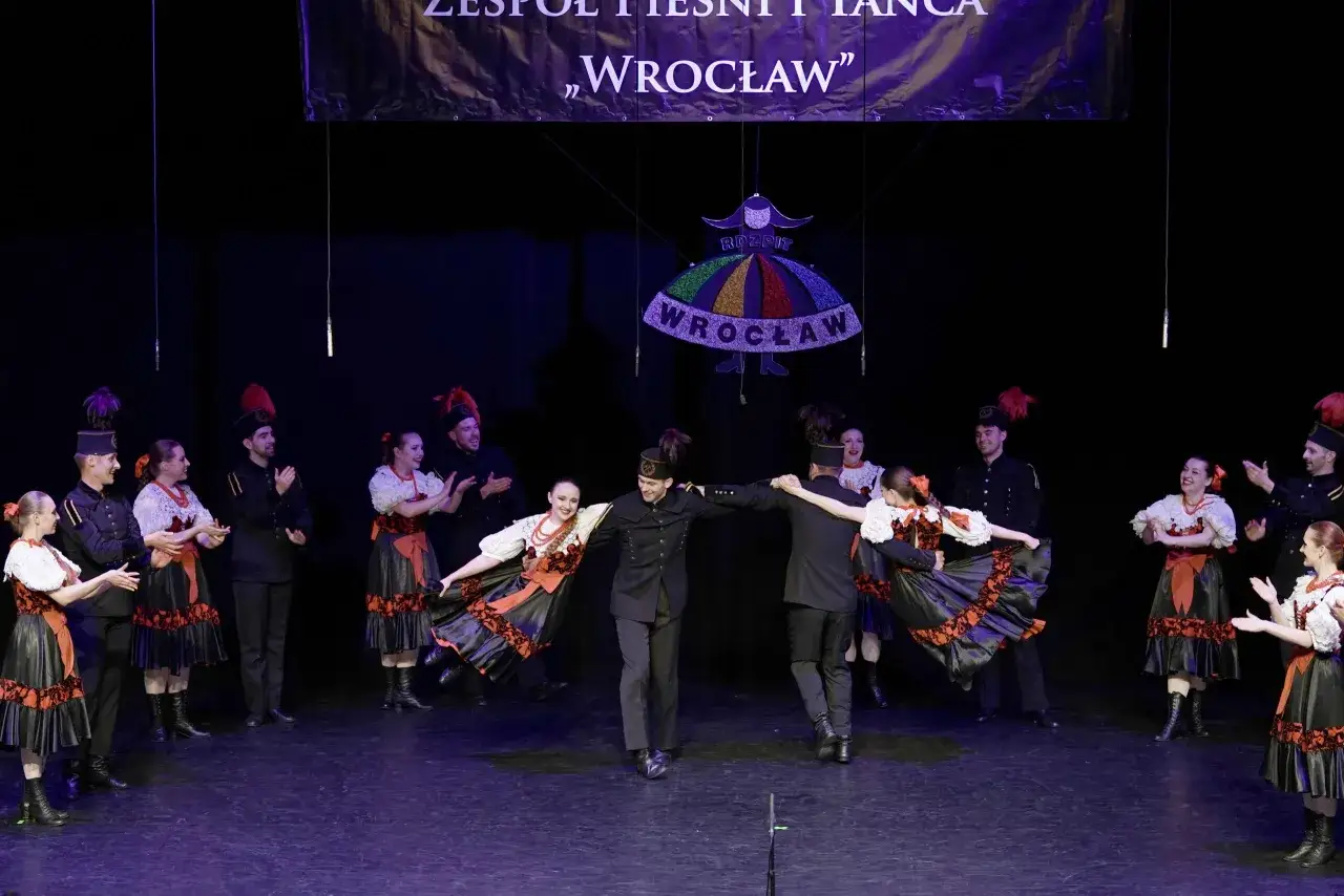 Tancerze w strojach ludowych wykonują taniec. Na scenie widać kolorowy baner z napisem
