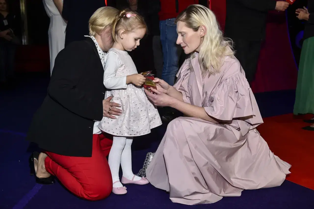 Agata Buzek z dziećmi, uśmiechnięta, wręcza prezent małej dziewczynce.