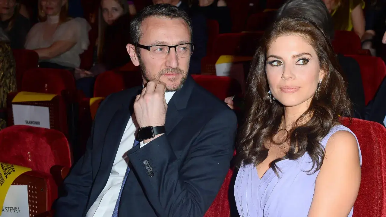 Weronika Rosati w liliowej sukni i jej partner, mężczyzna w okularach, siedzą obok siebie w teatrze.
