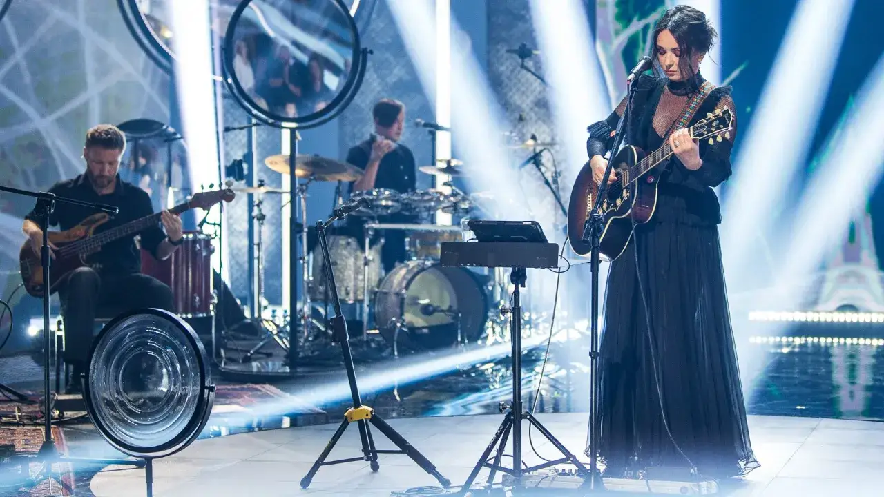 Kasia Kowalska na scenie z zespołem, grając na gitarze. W tle perkusja i basista.