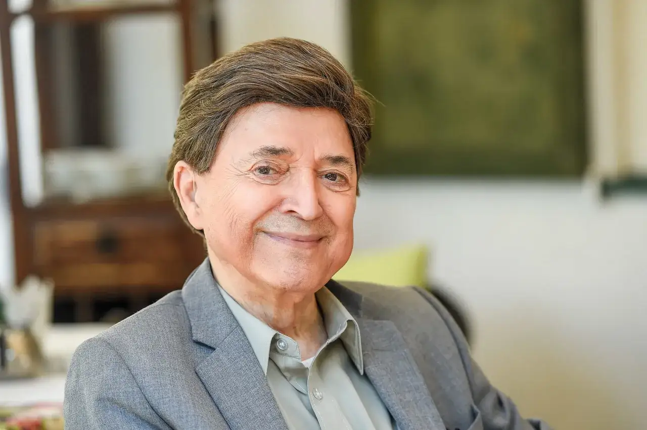 Uśmiechnięty Jerzy Połomski w szarym garniturze. W tle widać fragmenty mebli i zieloną ścianę.